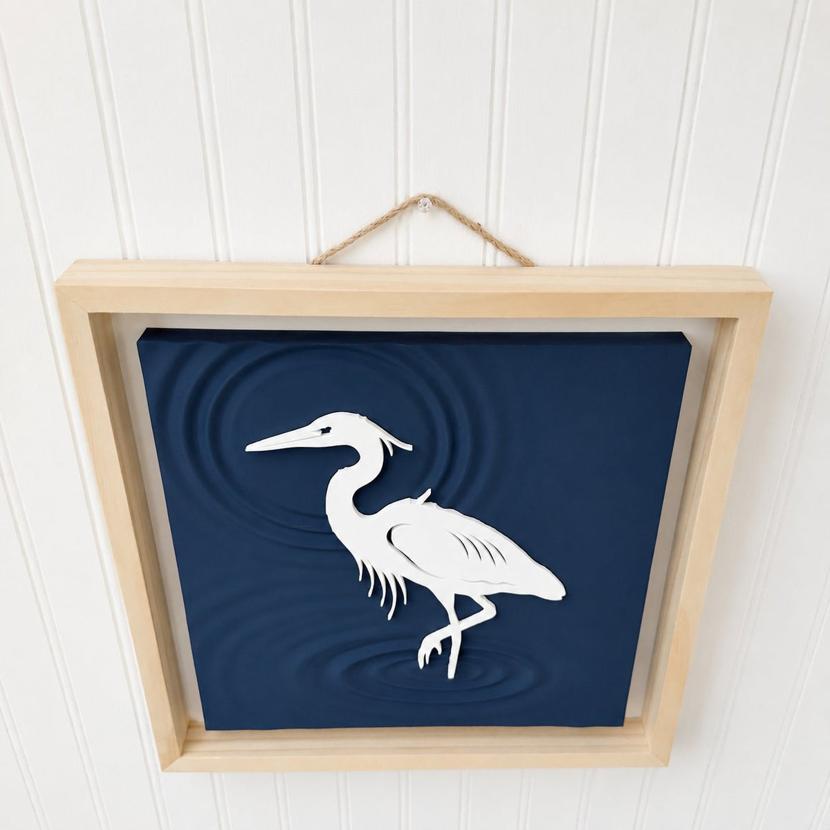 Framed heron wall art hanging by a jute rope on a white shiplap wall, viewed from below